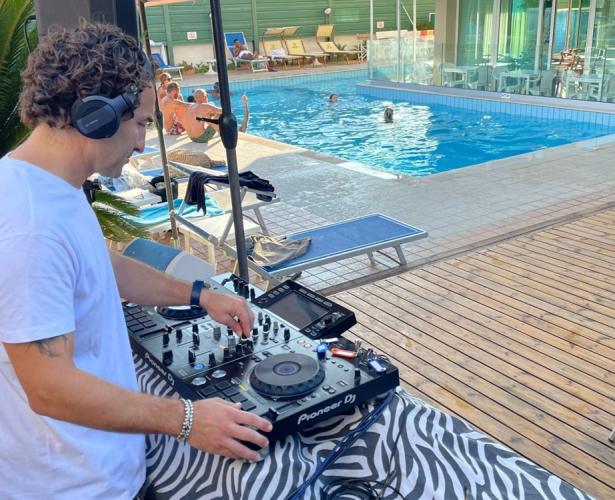 DJ suona musica a bordo piscina, persone si rilassano e nuotano.