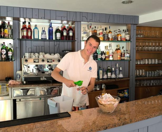 Barista prepara un drink dietro il bancone di un bar ben fornito.