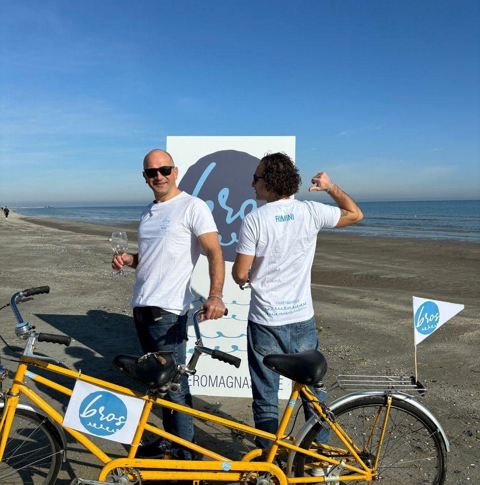 Due uomini con magliette bianche posano sulla spiaggia con una bicicletta gialla.